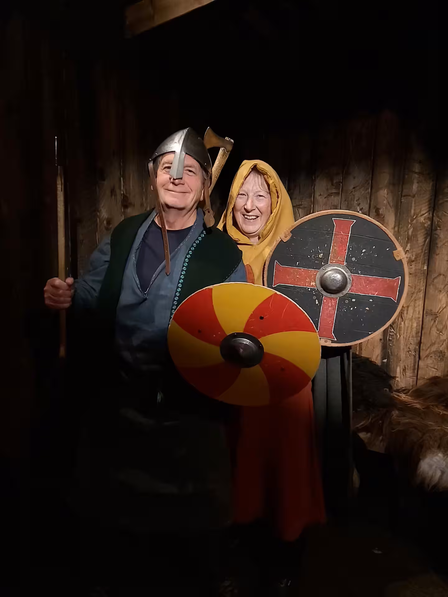 Two smiling visitors in Viking costumes holding round shields and spear inside a wooden longhouse in Skagafjordur