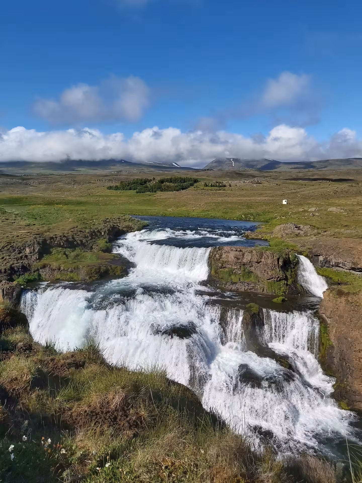 Cascading waterfall over rocky ledges in Skagafjordur valley with green meadows and mountains beyond