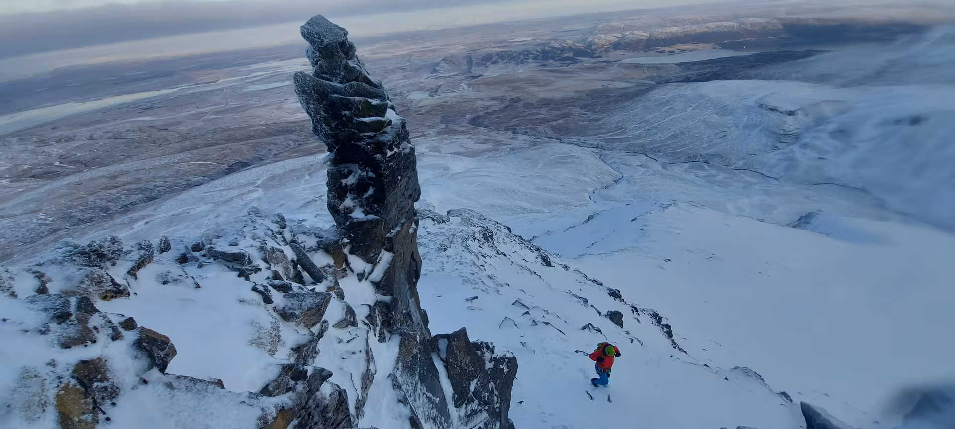 Climber ascending snow-covered Skessuhorn mountain ridge with dramatic rock pinnacle and vast arctic landscape beyond