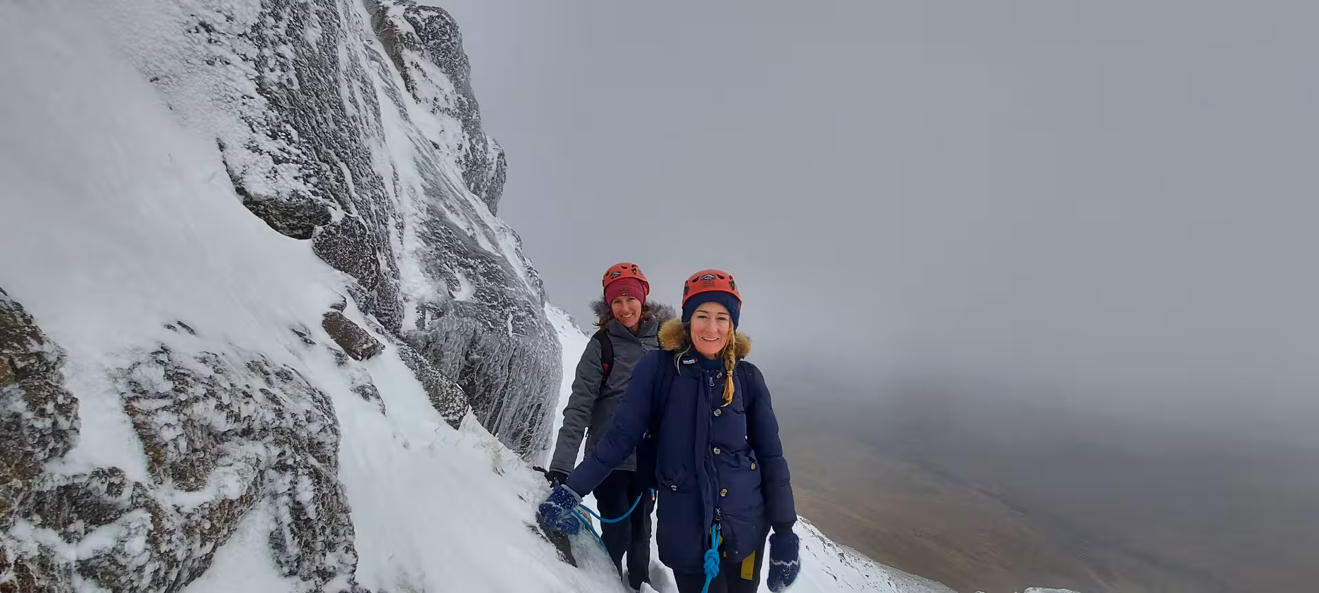 Two women in red helmets and winter gear climbing snowy Skessuhorn alpine mountain ridge in misty conditions