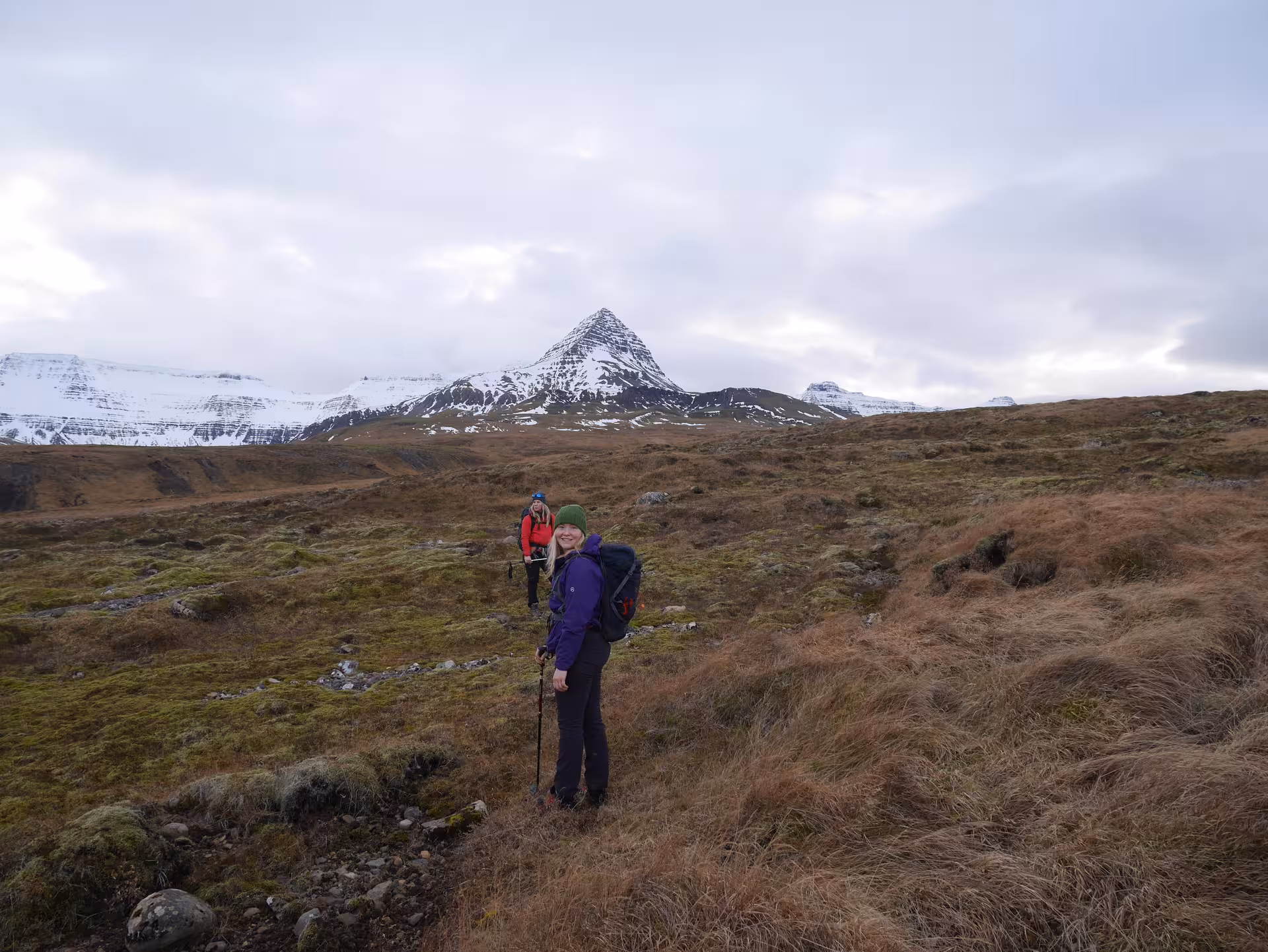 Two hikers trekking across alpine tundra with snow-capped Skessuhorn mountain peak in background, Iceland