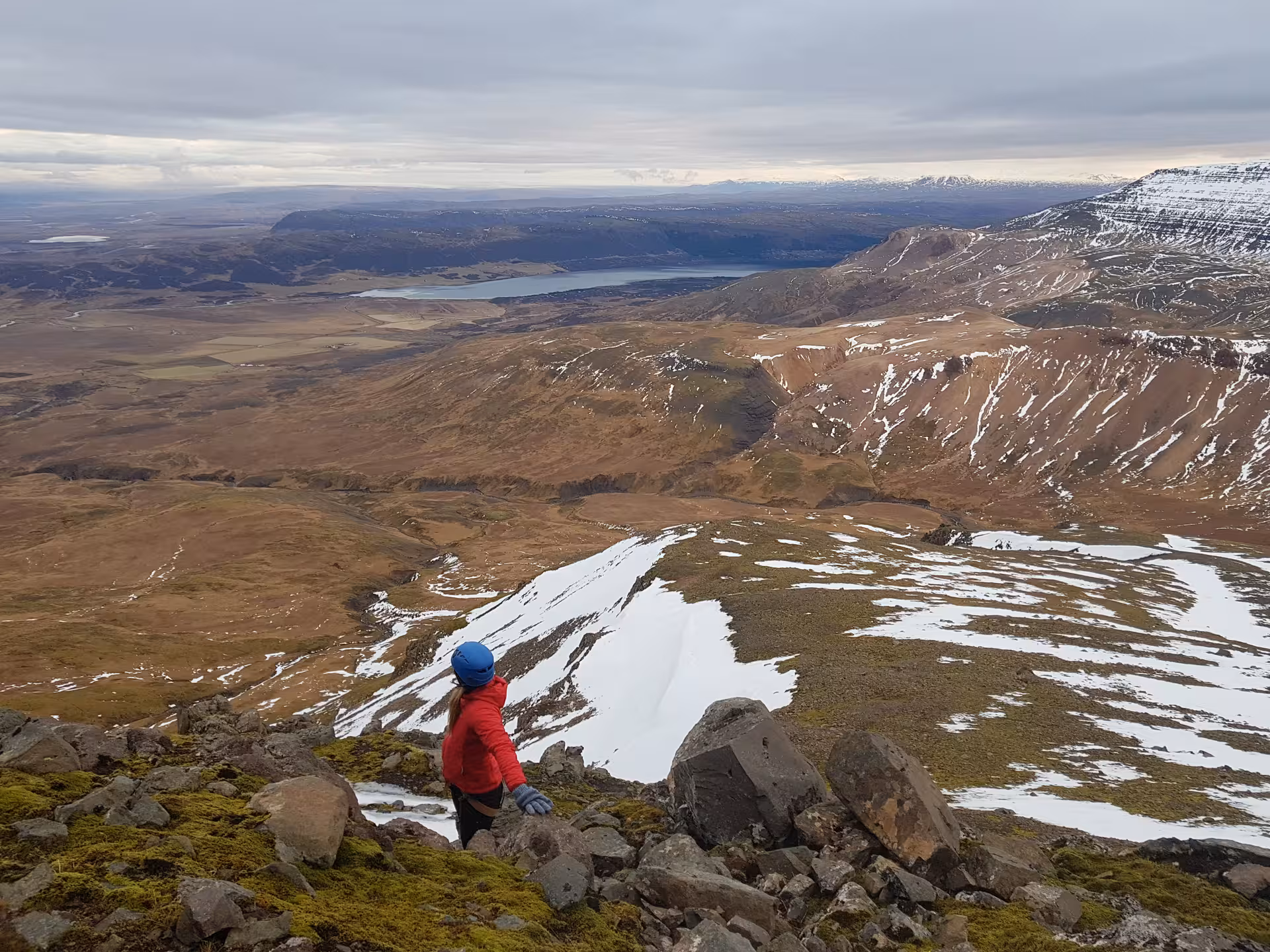 Hiker in red jacket overlooking vast Icelandic highland landscape with snow patches and lakes from Skessuhorn mountain summit