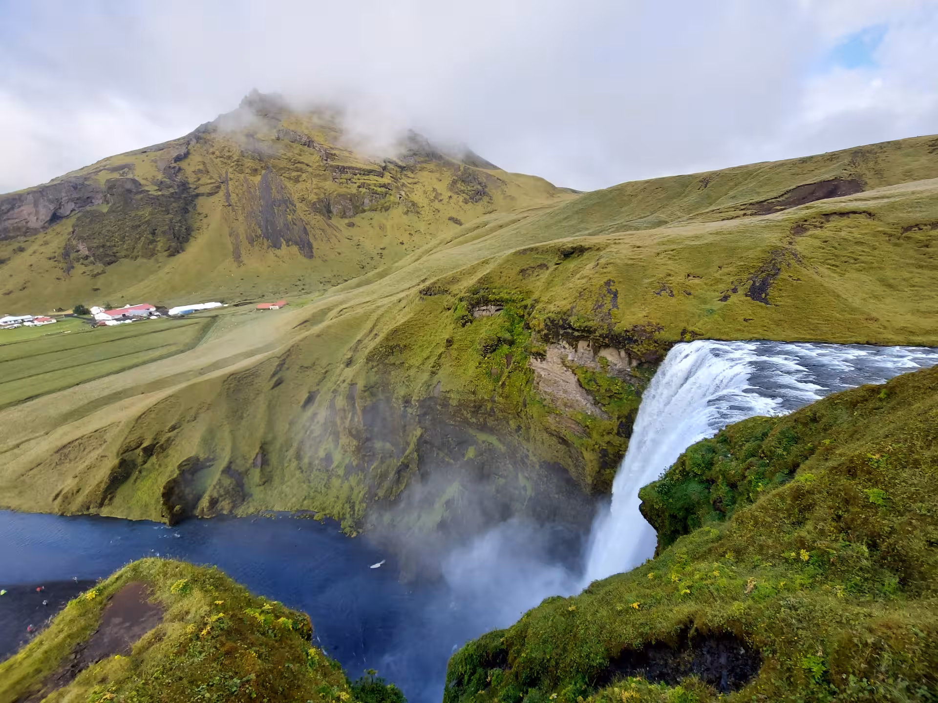 Skógafoss waterfall cascading over green cliffs with misty mountains and farmland in Iceland