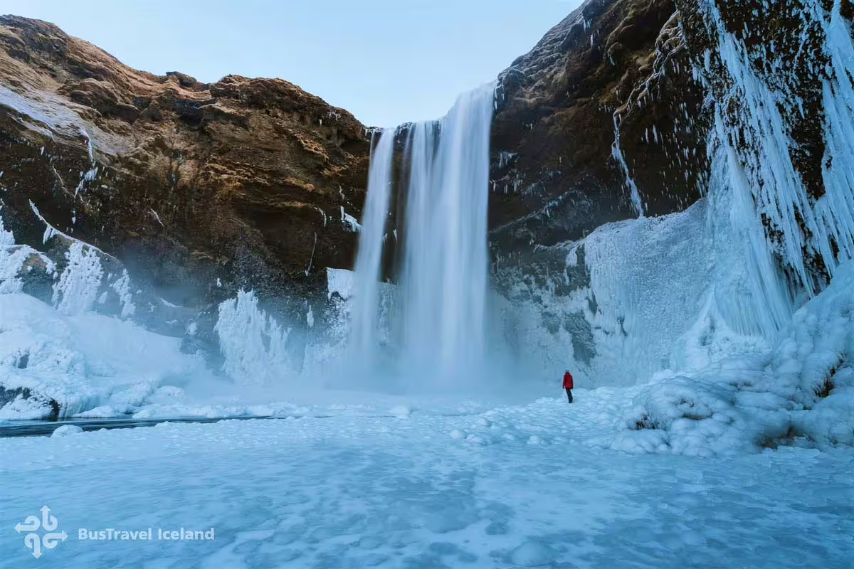 Person in red jacket standing before Skógafoss waterfall surrounded by ice and snow in Iceland winter