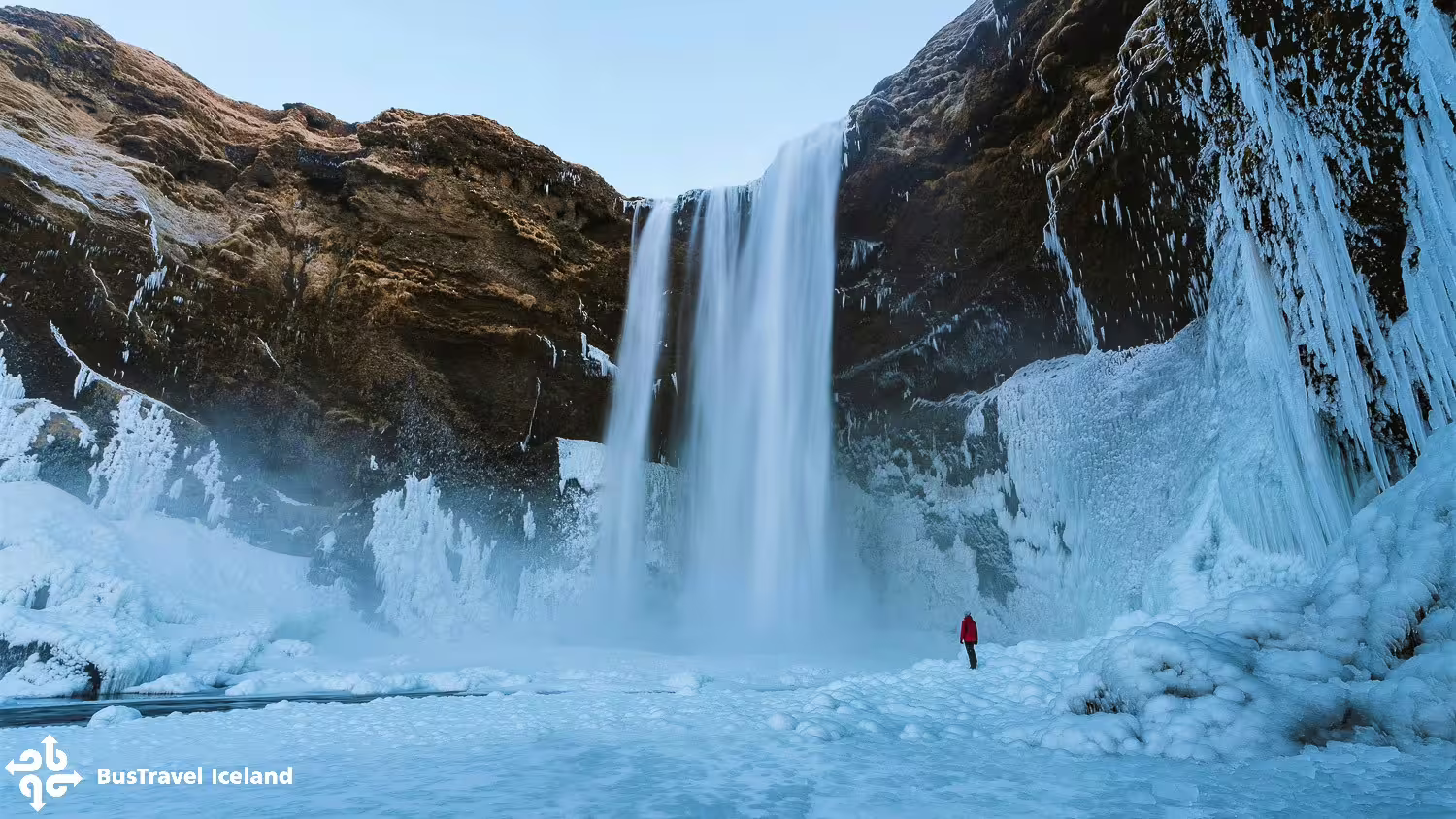 Person in red jacket standing before Skógafoss waterfall surrounded by ice and snow in Iceland winter