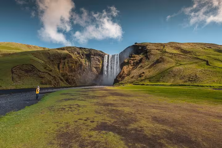 Skógafoss waterfall on Iceland's South Coast with a lone visitor in a yellow jacket standing on green plains