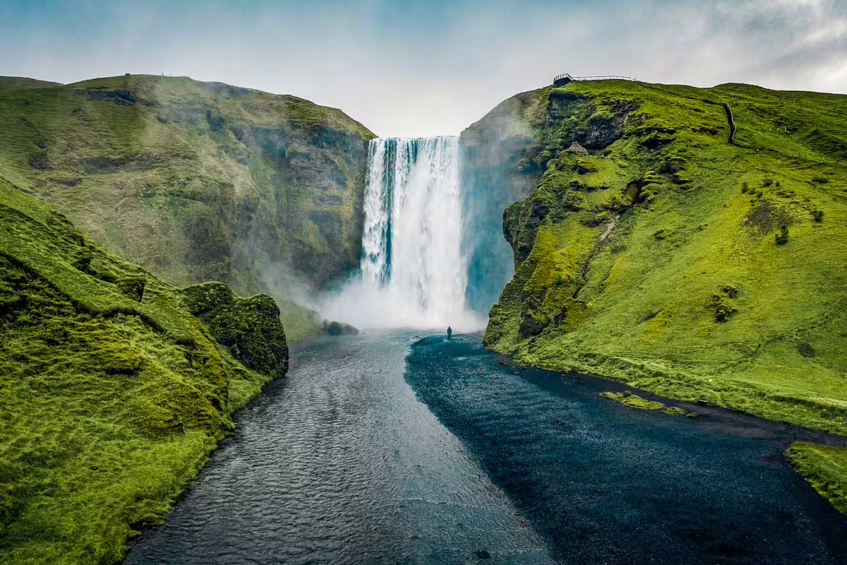 Skógafoss waterfall on Iceland's South Coast with a lone person standing at its base surrounded by green cliffs