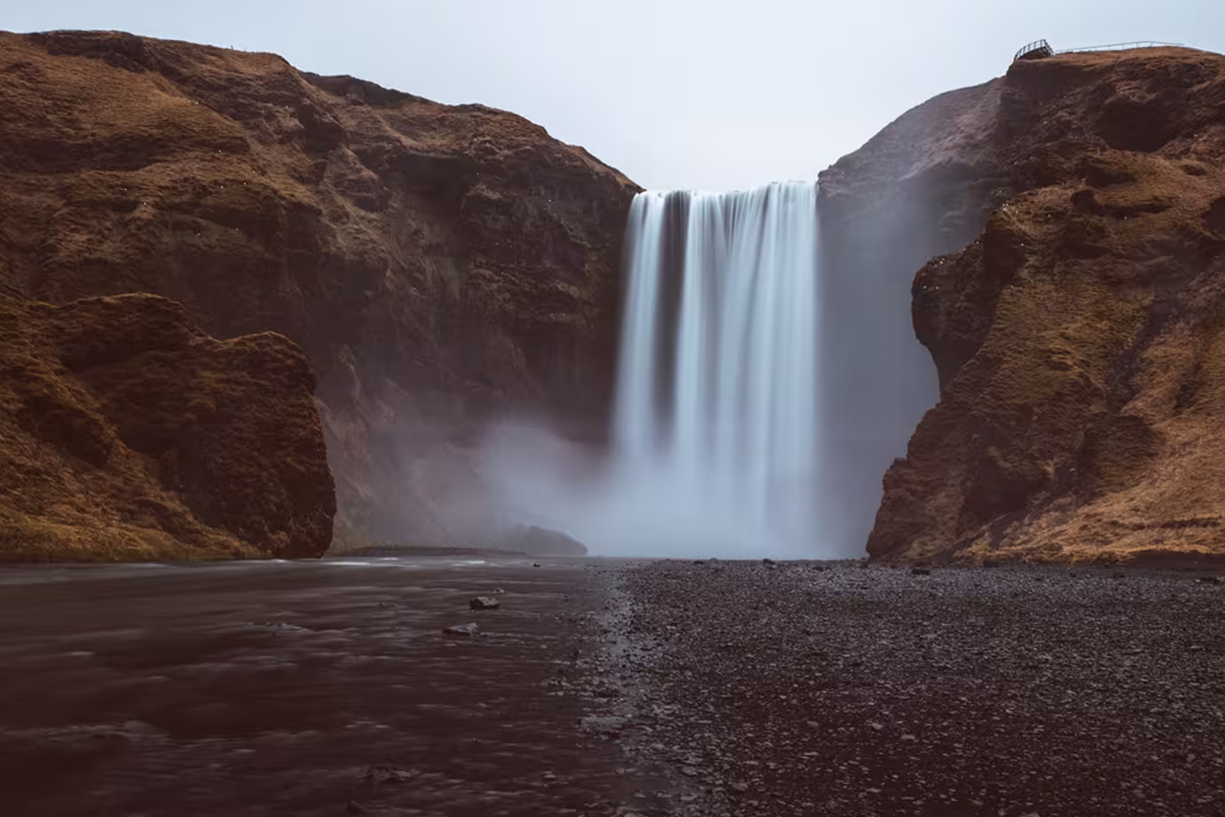 Skógafoss waterfall cascading down rocky cliffs on Iceland's South Coast with misty base and dark gravel riverbed