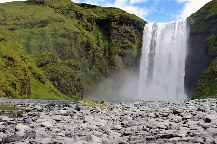 Massive Skógafoss waterfall cascading down mossy green cliffs onto rocky riverbed on Iceland's South Coast