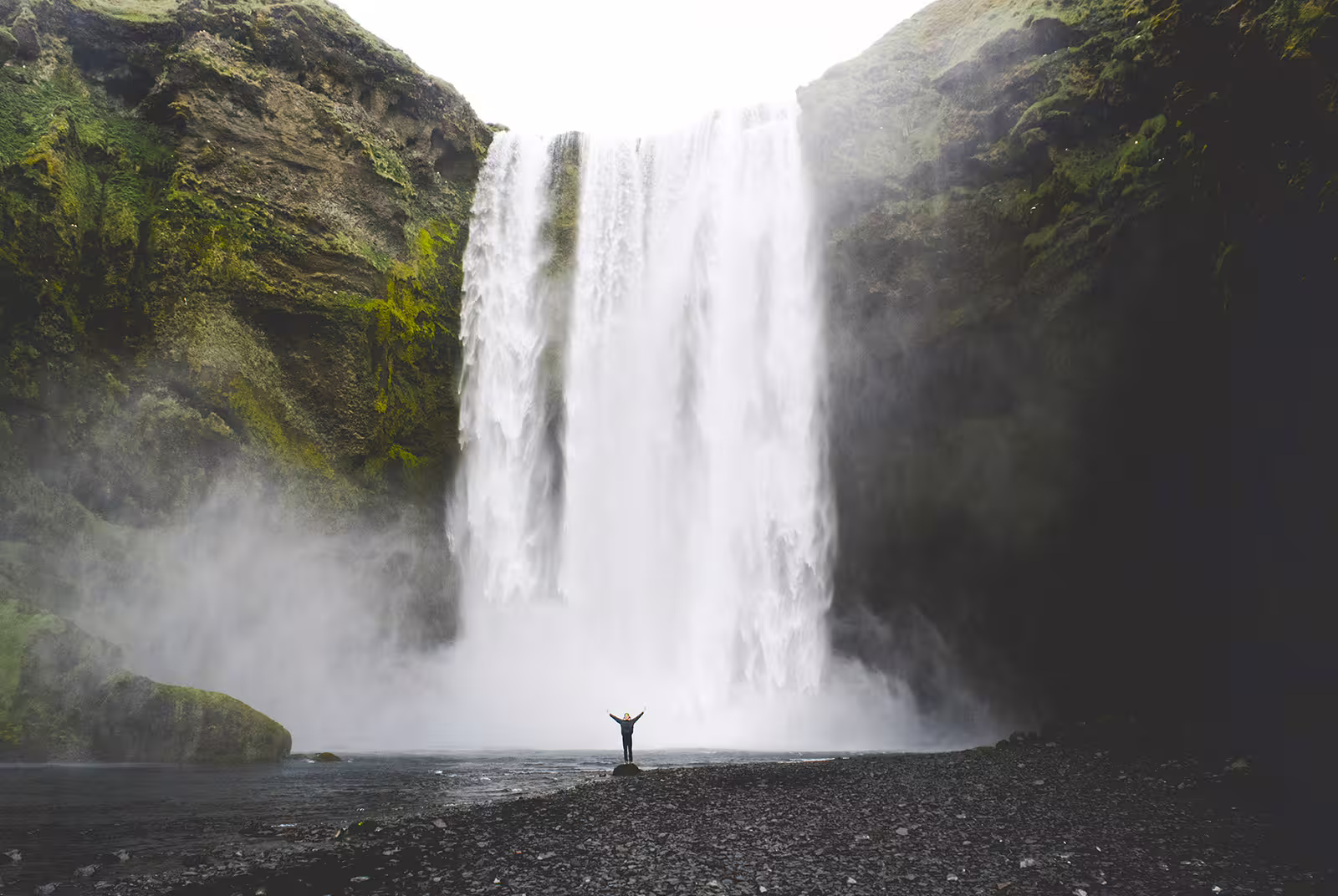 Person standing with arms raised in front of massive Skógafoss waterfall on Iceland's south coast