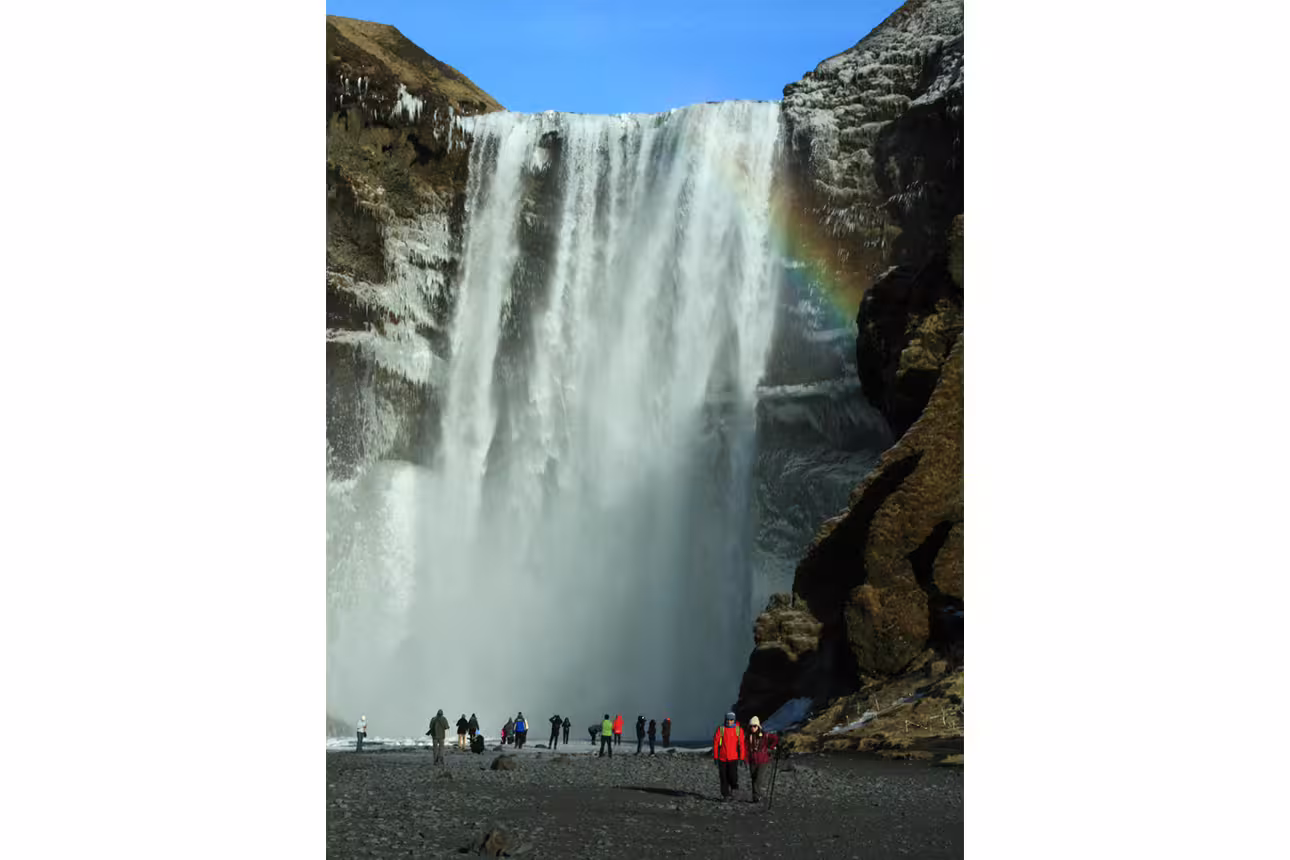 Tourists standing at the base of Skógafoss waterfall in Iceland with a rainbow visible in the mist