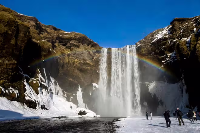 Skógafoss waterfall with rainbow and snow-covered ground, visitors standing in foreground, Iceland south coast