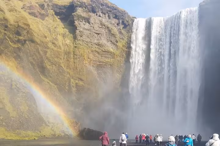 Tourists standing before Skógafoss waterfall with rainbow on Iceland South Coast tour