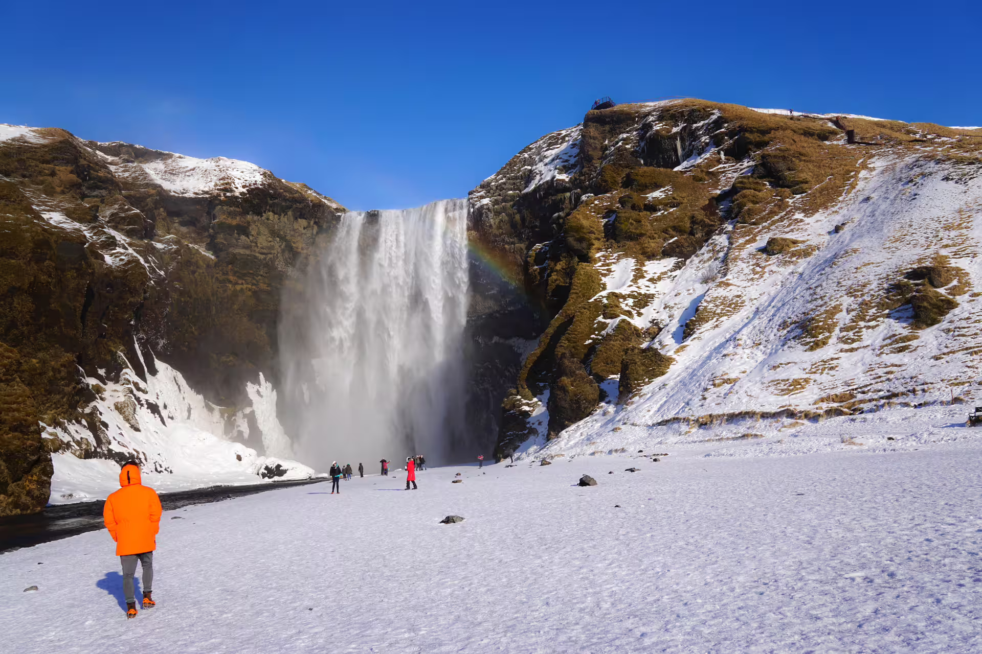 Tourist in orange jacket viewing Skógafoss waterfall with rainbow in snowy winter landscape, Iceland