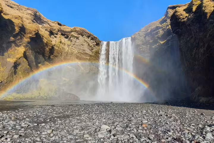 Skógafoss waterfall in South Iceland with a vibrant full rainbow arc over rocky ground under clear blue sky