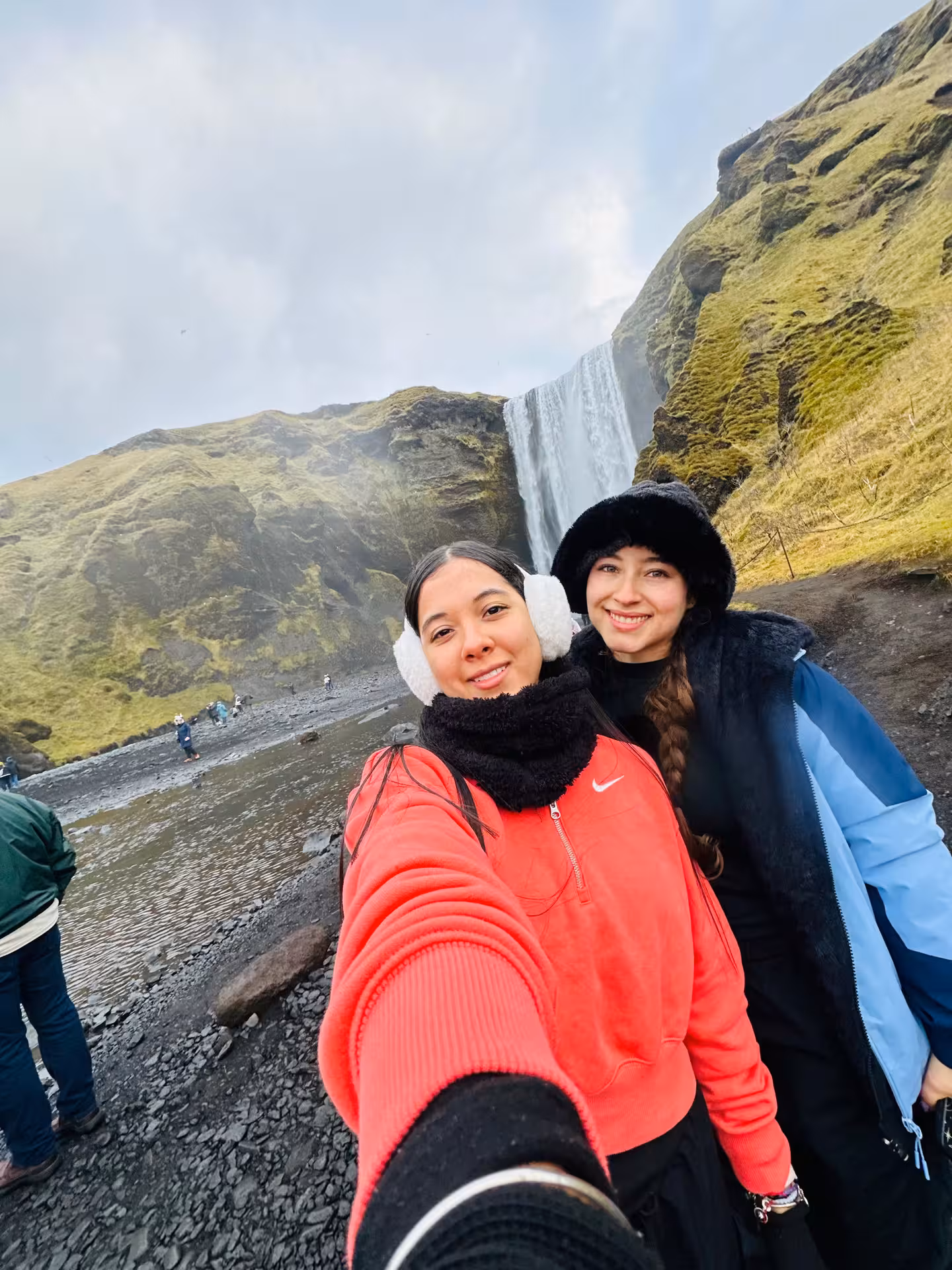 Two women smiling in front of Skógafoss waterfall in Iceland on a cloudy day