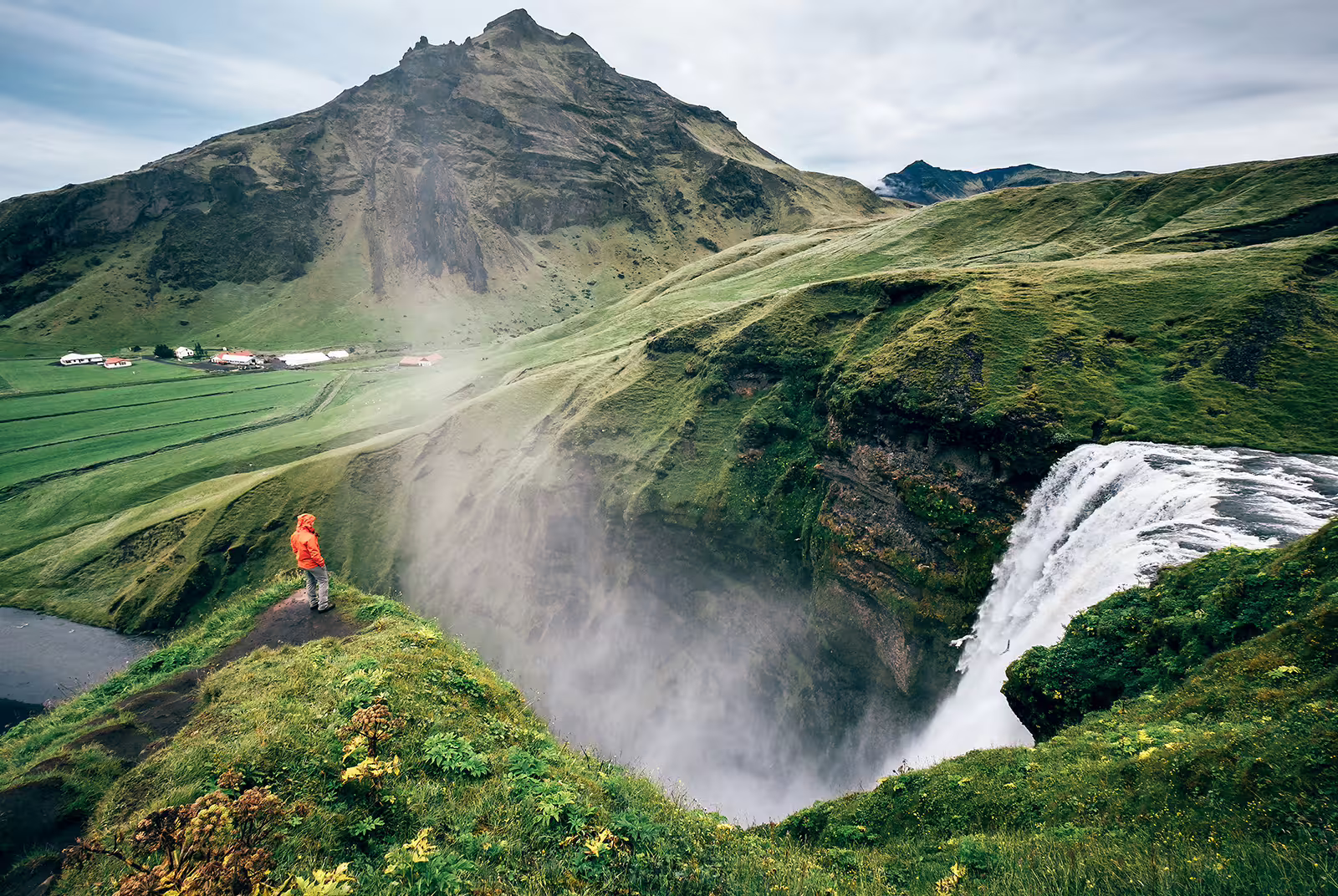 Hiker in red jacket standing at edge of Skógafoss waterfall with green Icelandic landscape and mountain behind