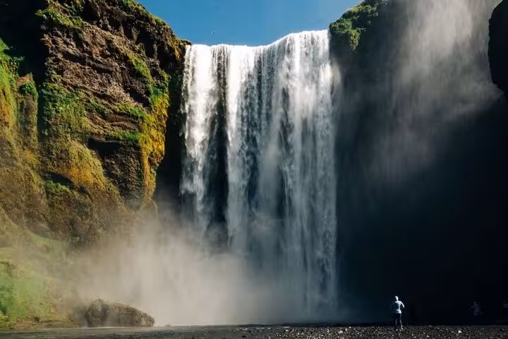Person standing before massive Skógafoss waterfall on Iceland's South Coast with mossy cliffs