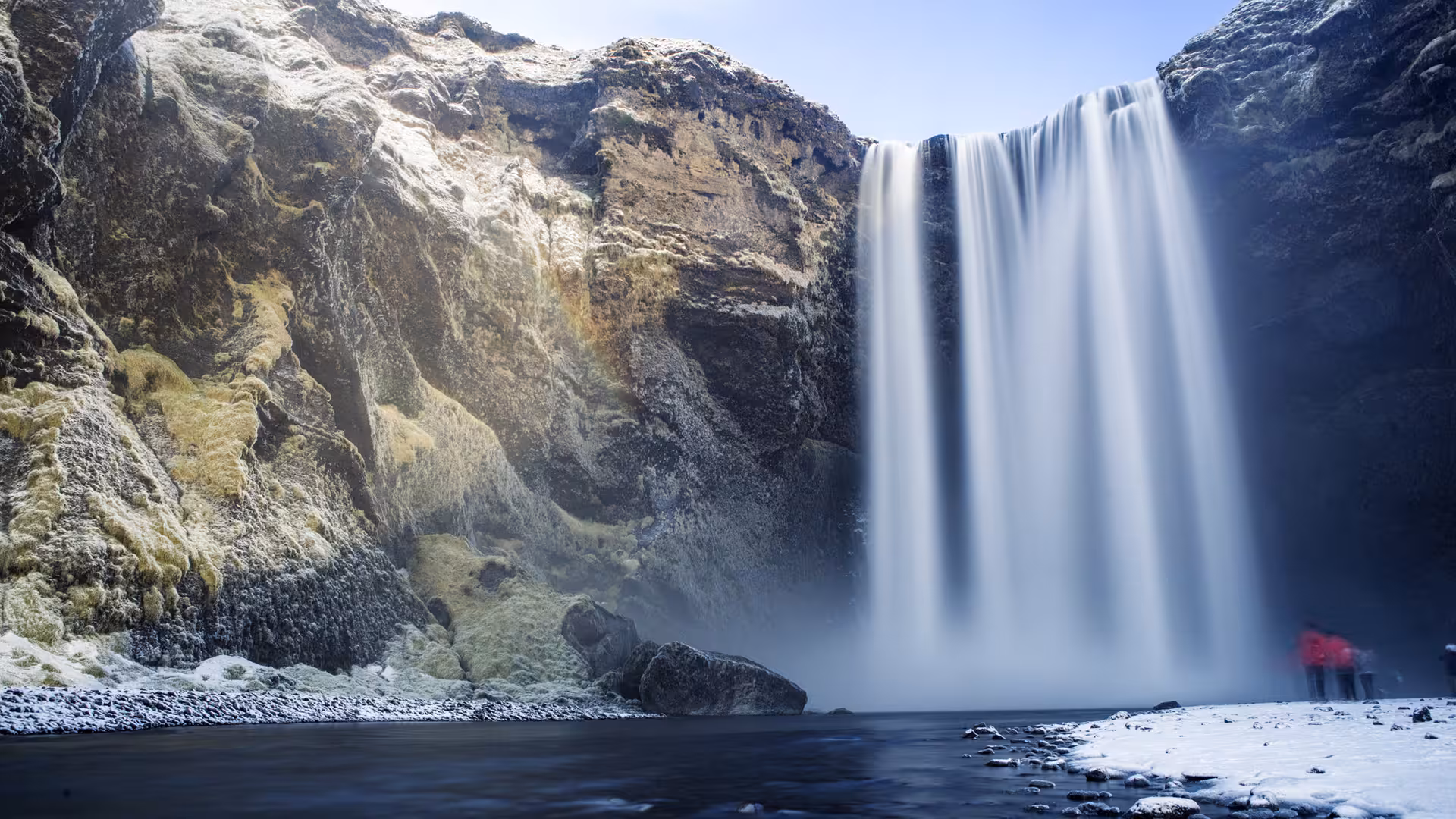 Skógafoss waterfall cascading down rocky cliffs in winter with visitors standing at its base, Iceland