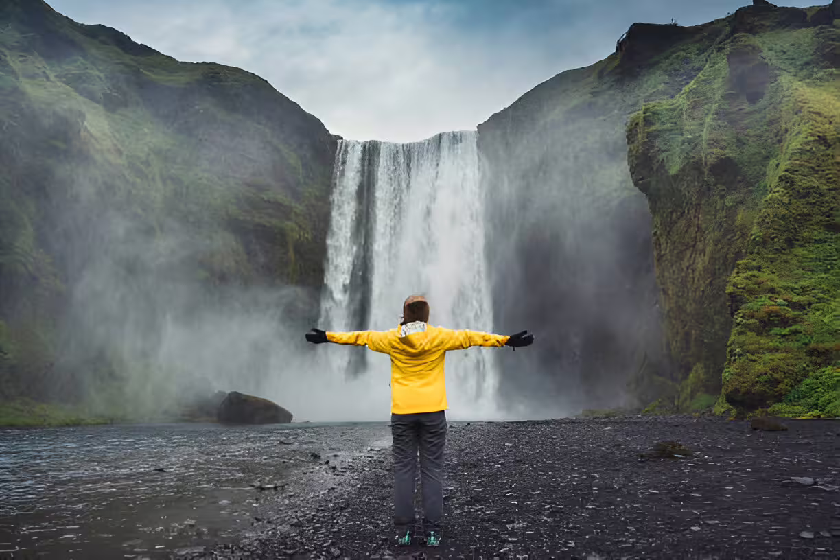 Traveler in yellow jacket standing arms wide open before Skógafoss waterfall on Iceland's south coast