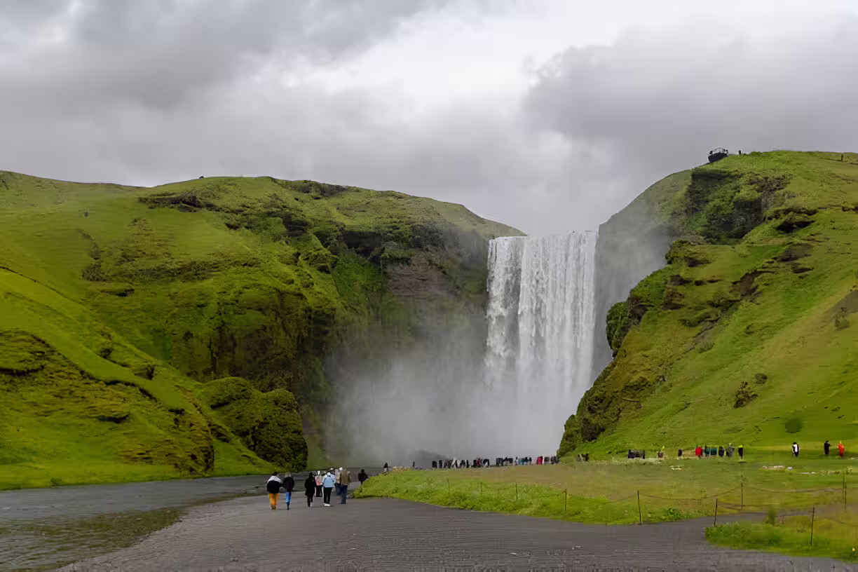 Tourists walking toward Skógafoss waterfall surrounded by green cliffs on Iceland's South Coast