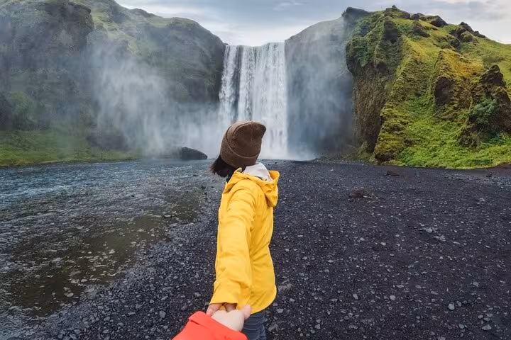 Traveler in yellow jacket holding hands facing Skógafoss waterfall on Iceland's black sand south coast