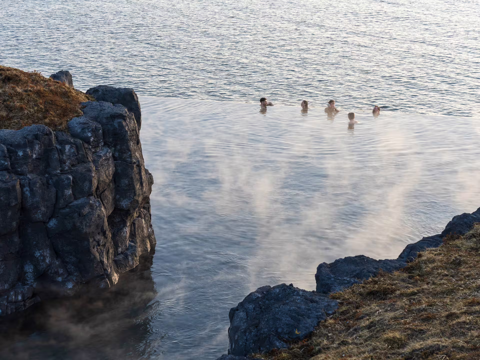 Group of people soaking in steaming Sky Lagoon geothermal pool by rocky coastline in Iceland