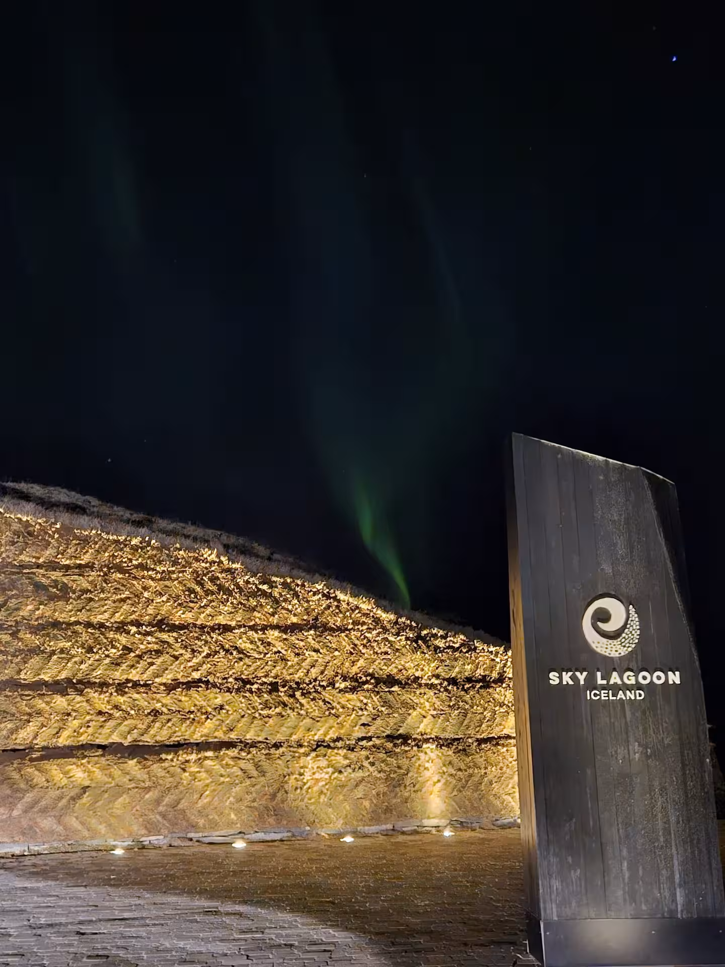 Sky Lagoon Iceland entrance sign with northern lights aurora borealis glowing green over illuminated lava wall at night