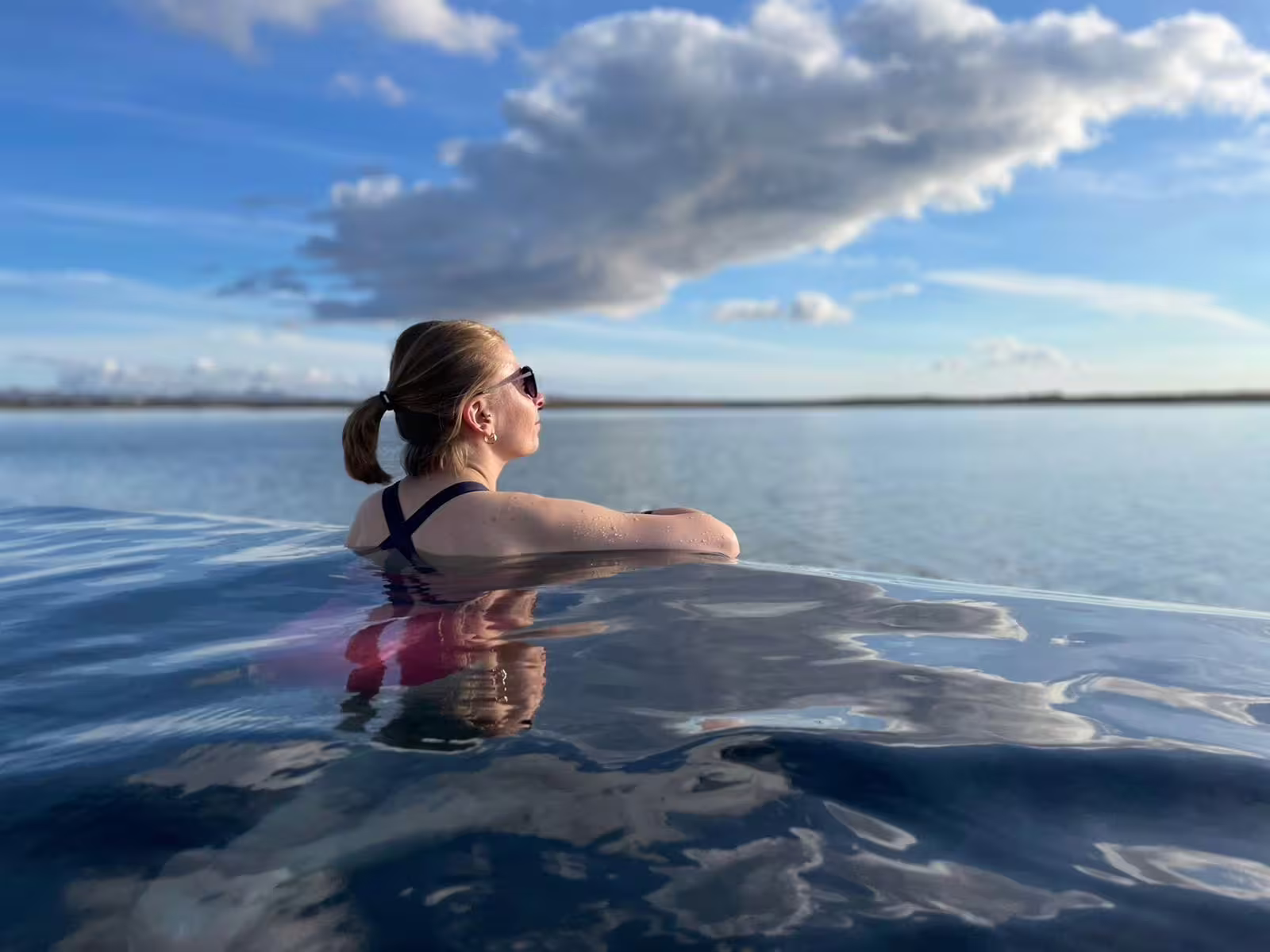 Woman relaxing in Sky Lagoon infinity pool overlooking calm Icelandic waters under a blue cloudy sky