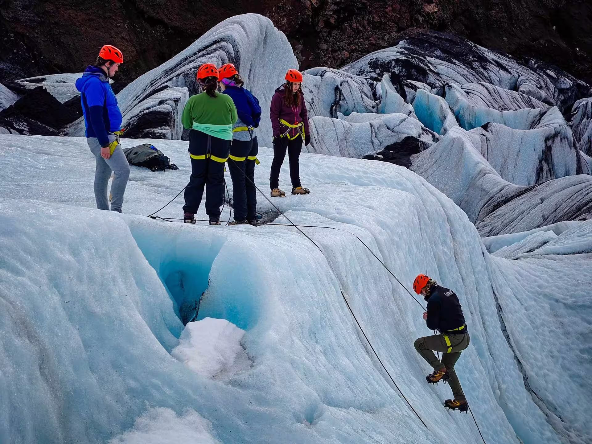 Group of hikers with orange helmets and harnesses on Solheimajokull glacier with guide rappelling ice face