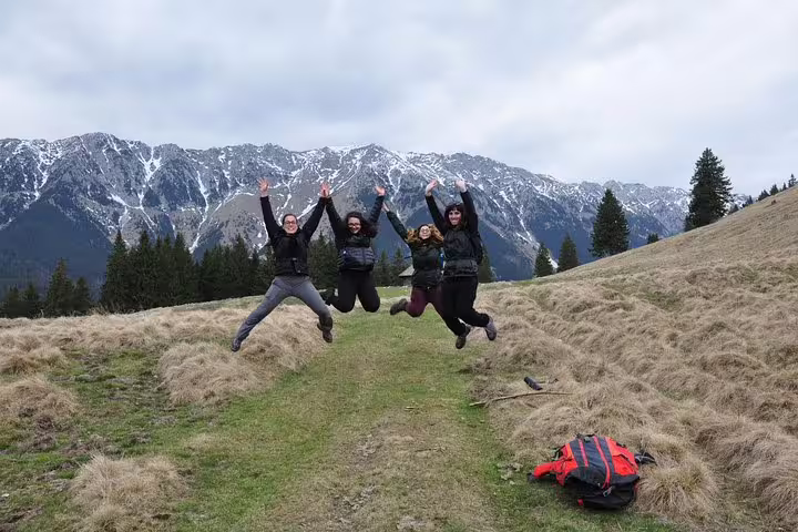 Four hikers jumping joyfully on a mountain trail with snow-capped Carpathian peaks near Brasov
