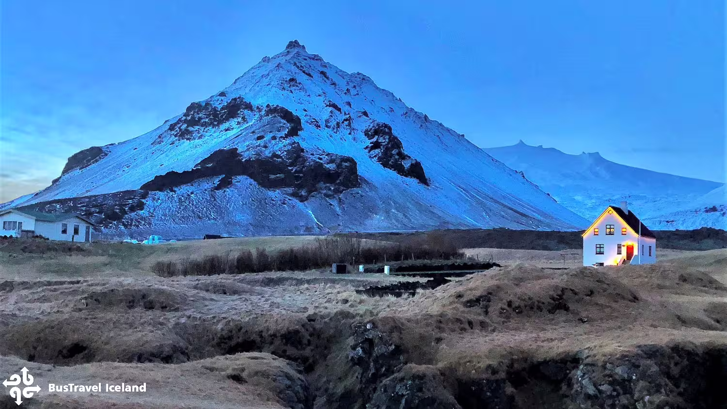 Snow-capped Snæfellsjökull volcano at dusk with a lit white farmhouse on Snæfellsnes Peninsula, Iceland