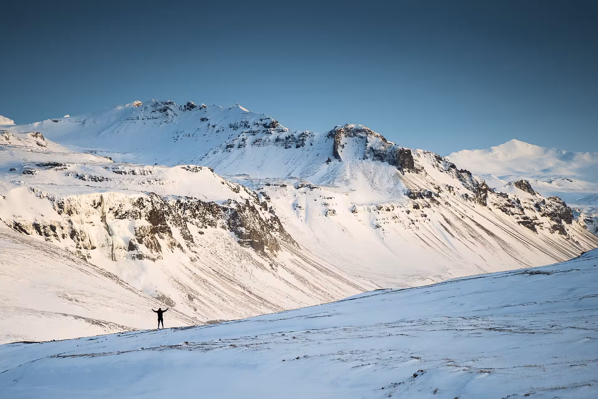 Person standing arms raised on snowy Snaefellsnes landscape with dramatic snow-covered volcanic mountains behind