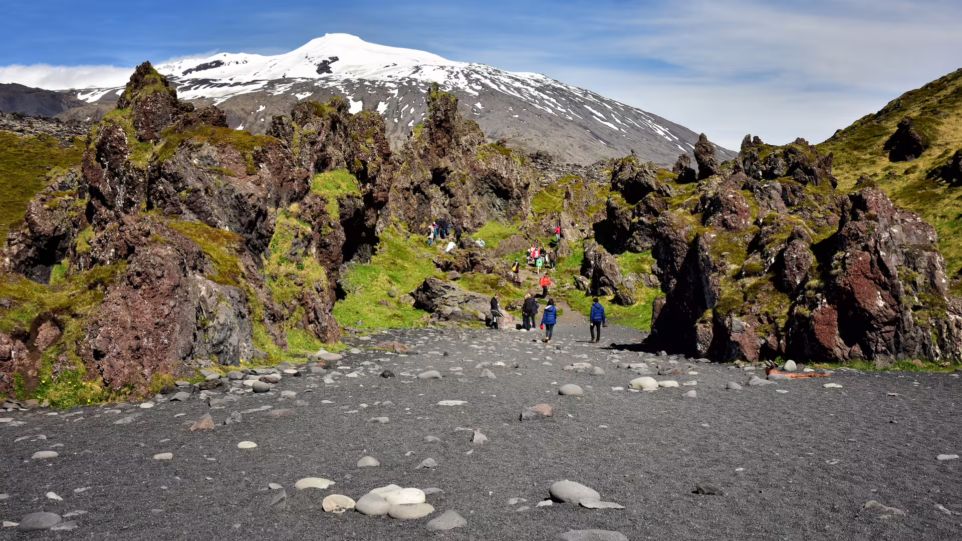 Hikers walking across black sand beach toward jagged lava rocks with snow-capped Snæfellsjökull glacier volcano behind