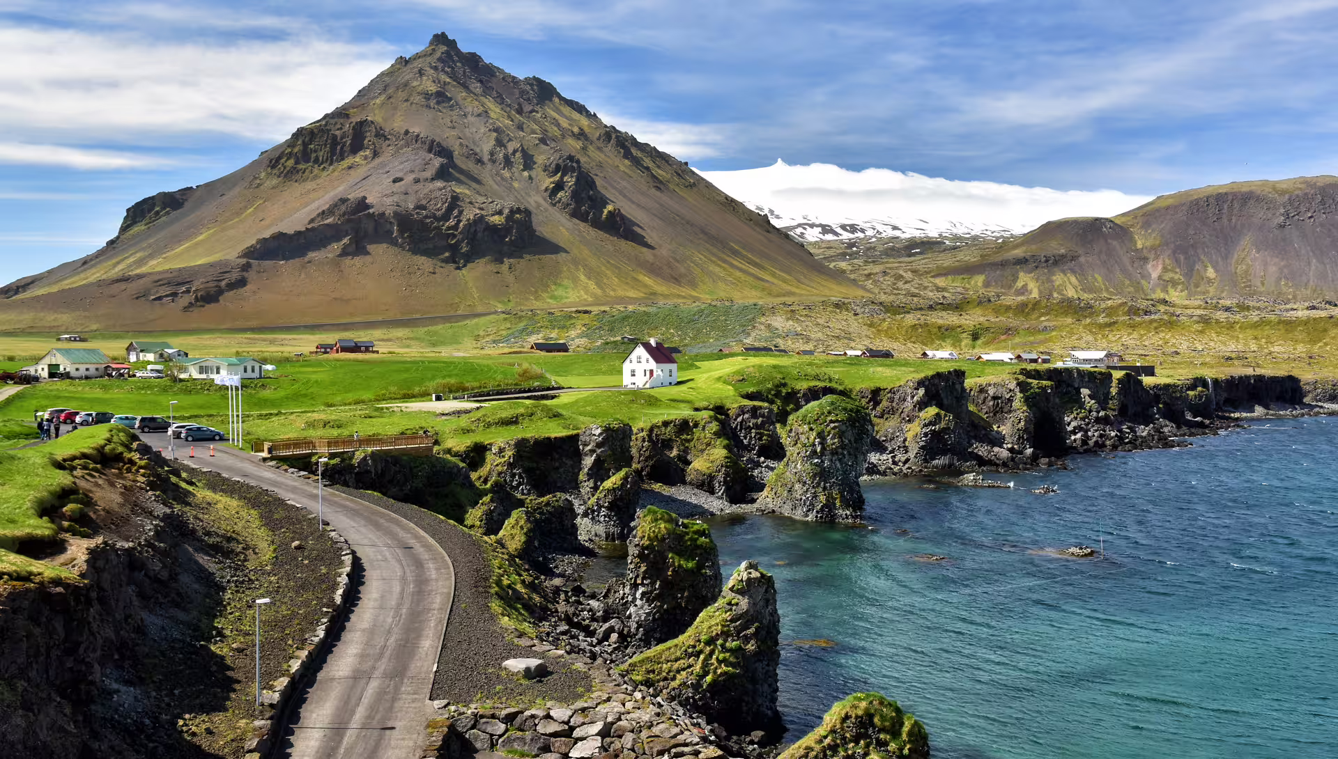 Coastal road winding past mossy lava cliffs toward green village with dramatic Snaefellsnes glacier volcano backdrop