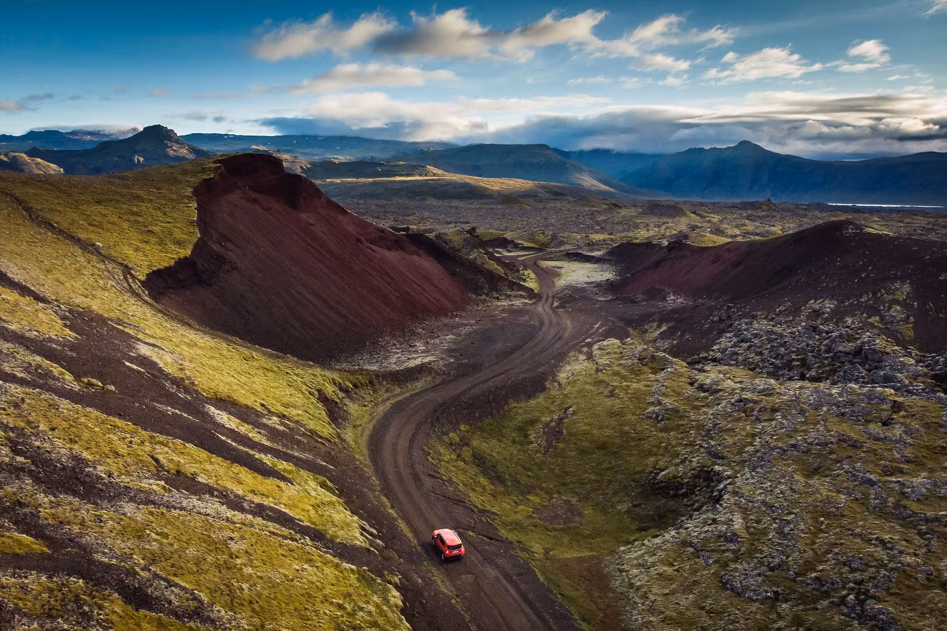 Red SUV driving on a winding dirt road through volcanic lava fields and green moss-covered hills in Iceland