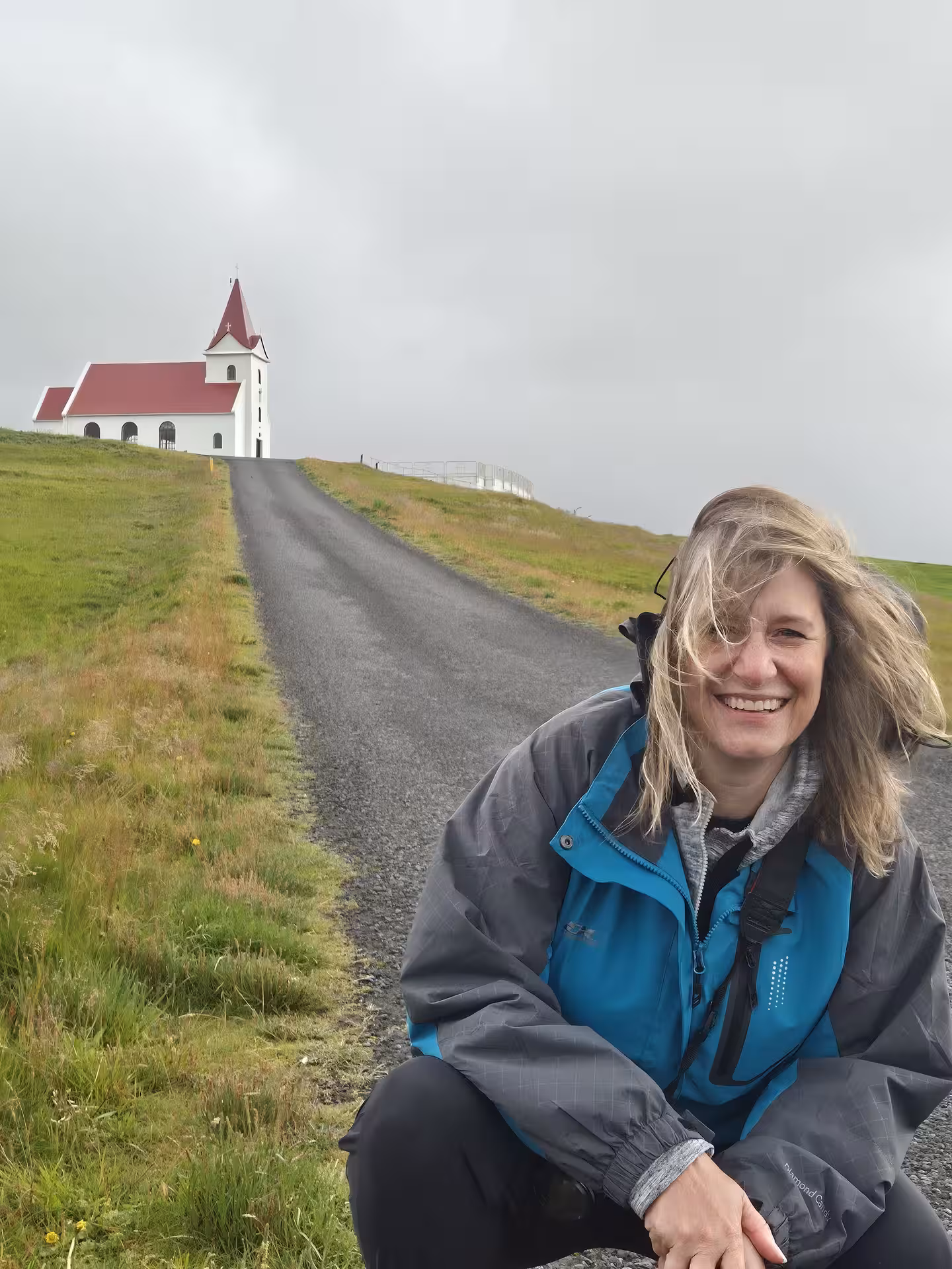 Smiling woman sitting on road leading to white red-roofed Icelandic church on Snaefellsnes Peninsula
