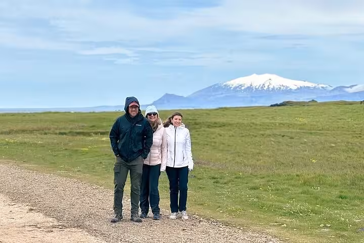 Three visitors posing on a gravel path with Snæfellsjökull glacier volcano and green fields in the background