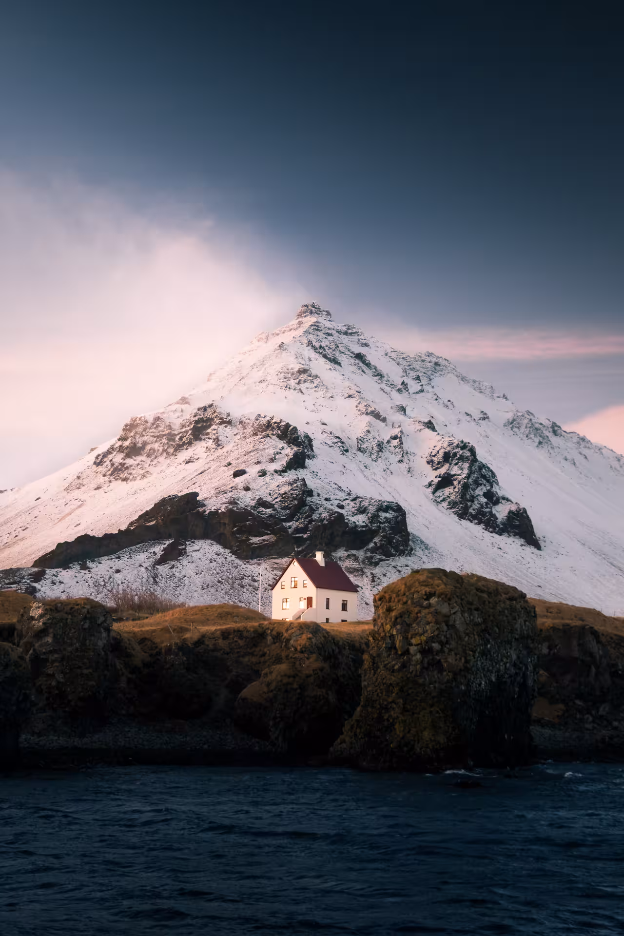White house with red roof beside rocky coast with snow-capped Snæfellsjökull mountain behind