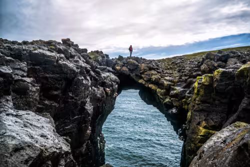 Hiker standing on natural rock arch over turquoise sea on Snaefellsnes Peninsula, Iceland