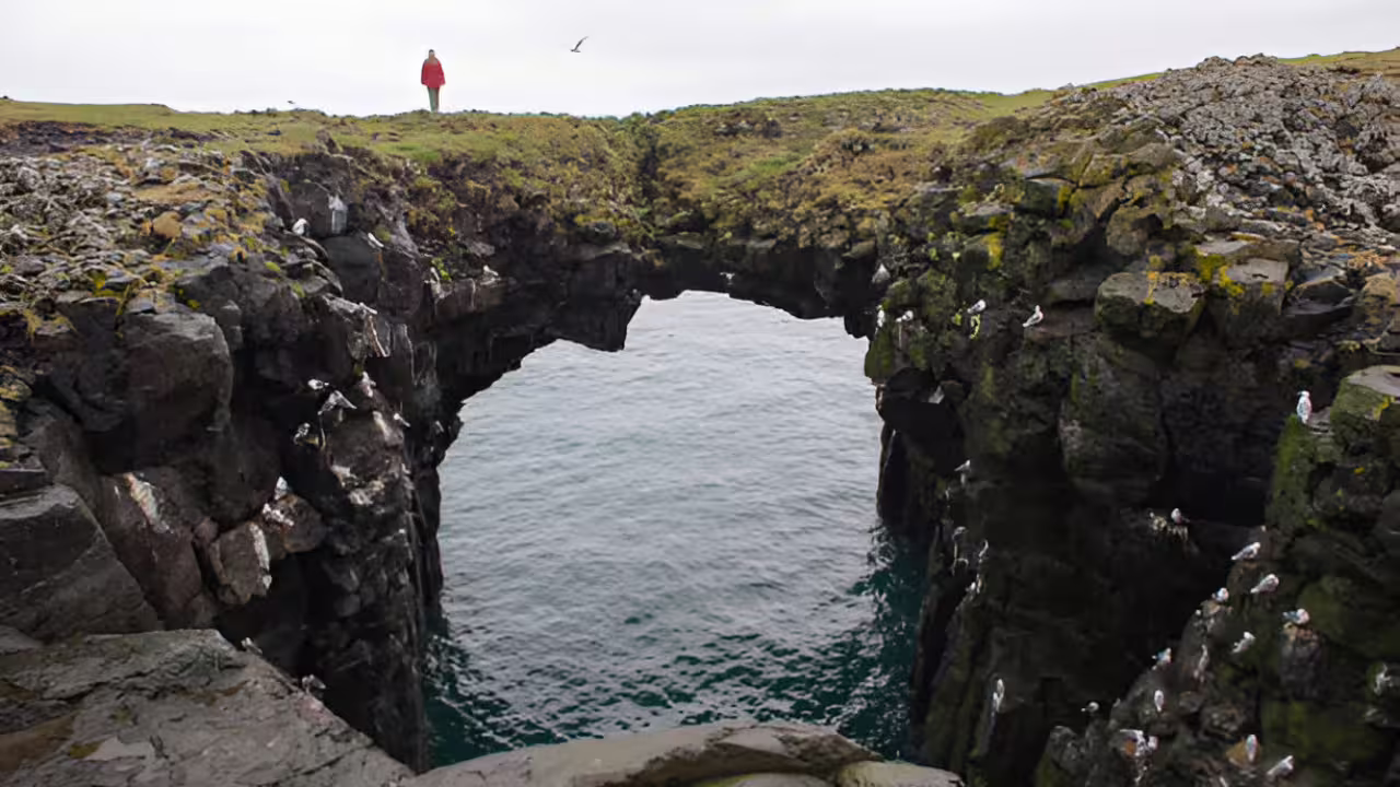 Natural rock arch over turquoise water on Snaefellsnes Peninsula with person in red jacket standing above