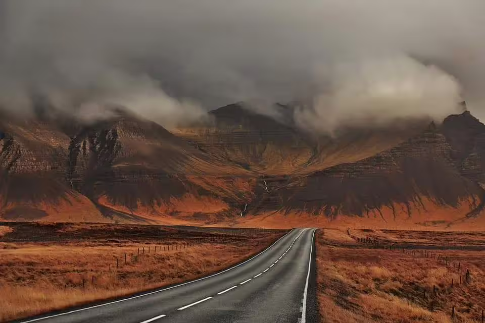 Two-lane road leading toward dramatic misty mountains with rust-colored moorland on Snaefellsnes Peninsula, Iceland