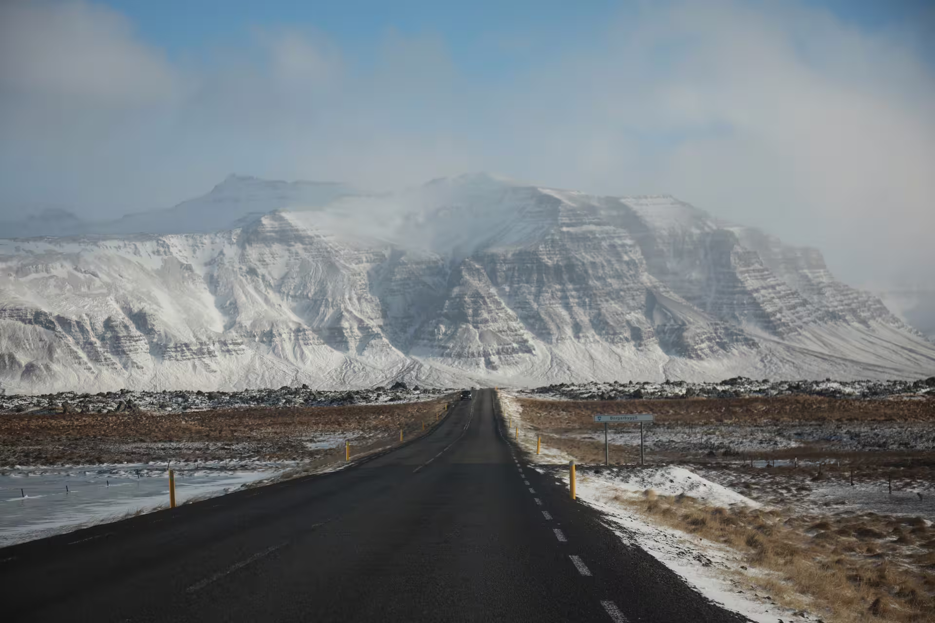 Two-lane road leading toward snow-covered layered mountains on Snaefellsnes Peninsula in winter