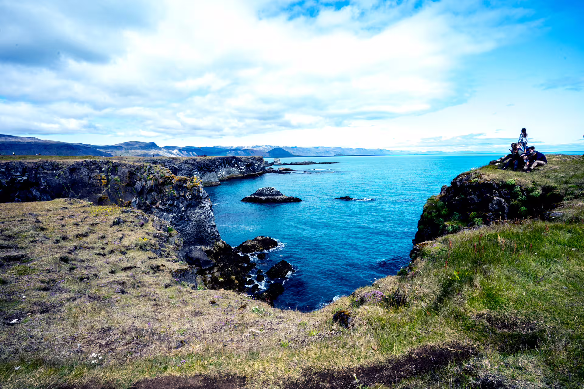 Rocky cliffs and vivid blue ocean cove on the Snaefellsnes Peninsula, Iceland, with visitors resting nearby
