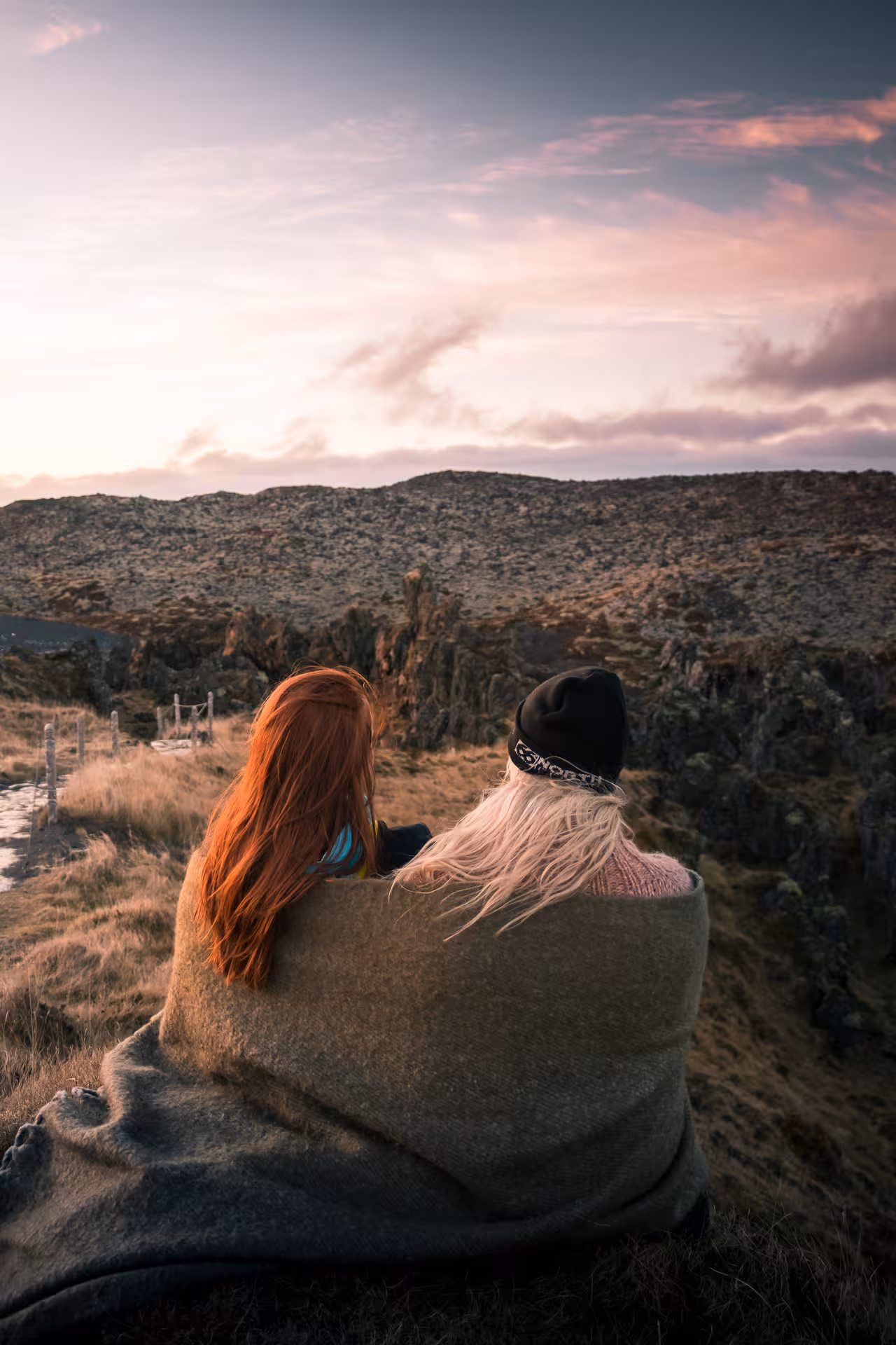 Two women wrapped in a blanket watching sunset over Snaefellsnes Peninsula rocky landscape
