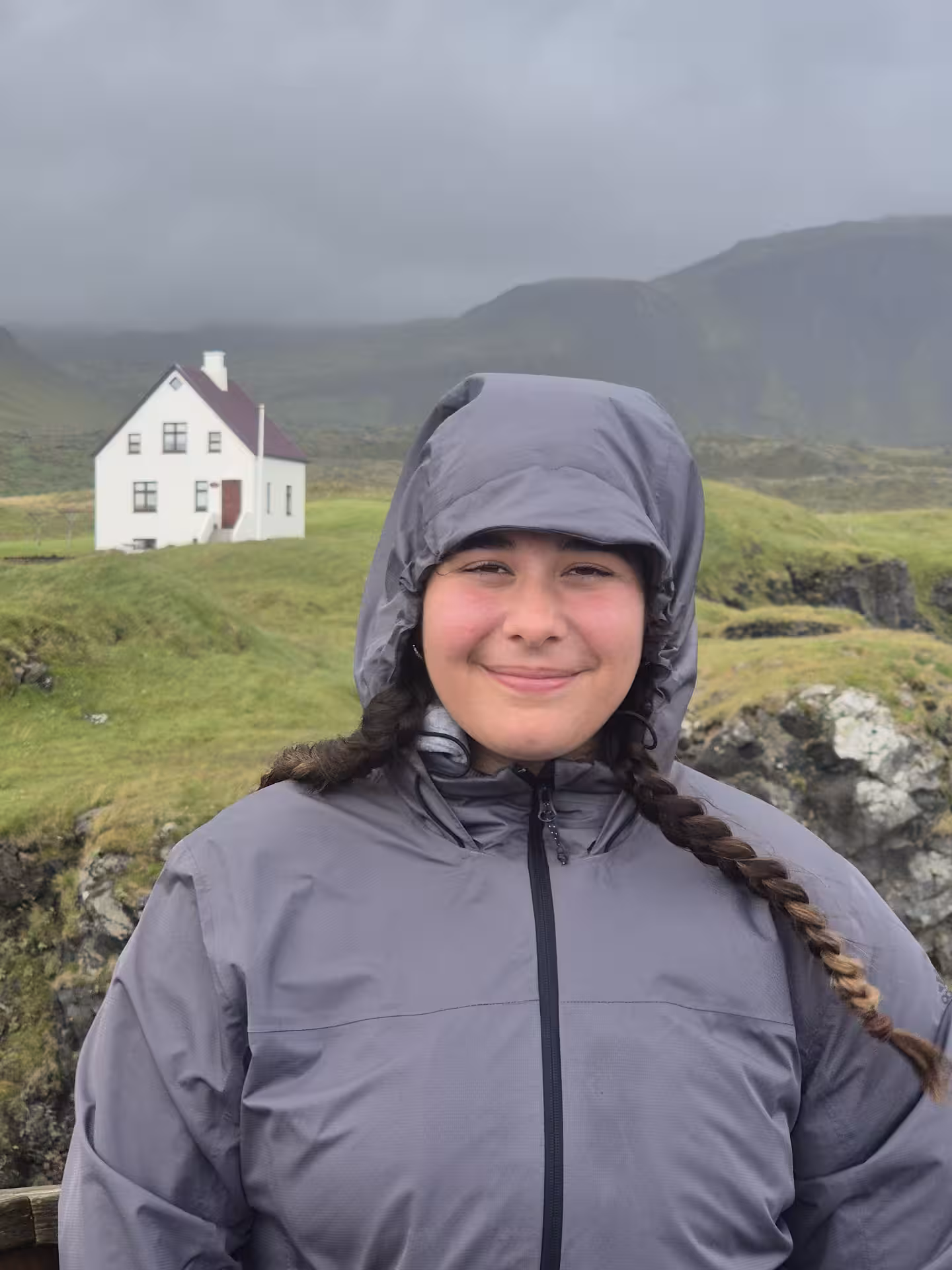 Smiling girl in gray rain jacket with braids standing before white farmhouse on green Snaefellsnes Peninsula