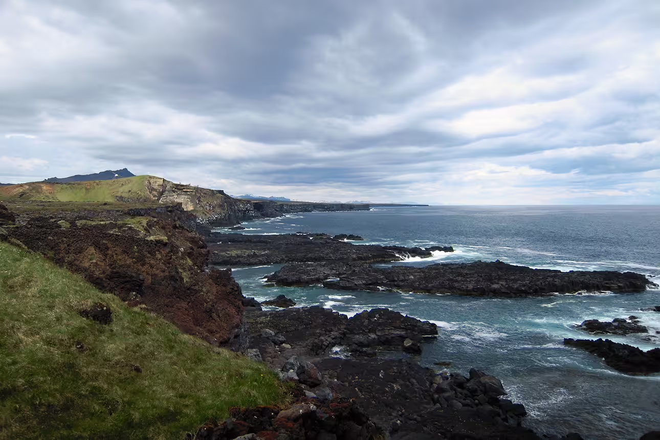 Rocky volcanic coastline of Snaefellsnes Peninsula with green cliffs and ocean waves under cloudy sky
