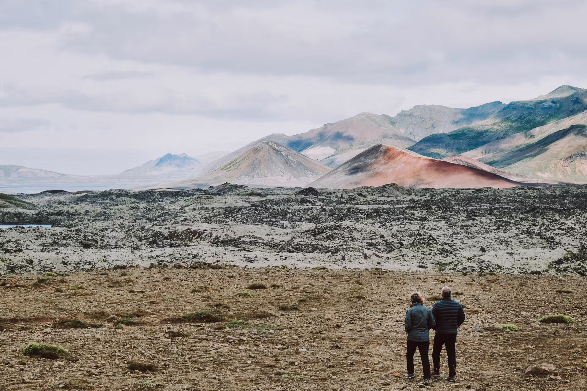 Two travelers stand before colorful volcanic mountains and lava fields on Snaefellsnes Peninsula, Iceland