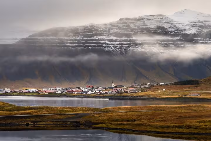 Small Icelandic coastal town with colorful rooftops beside a fjord, dramatic snow-capped mountains and low clouds behind