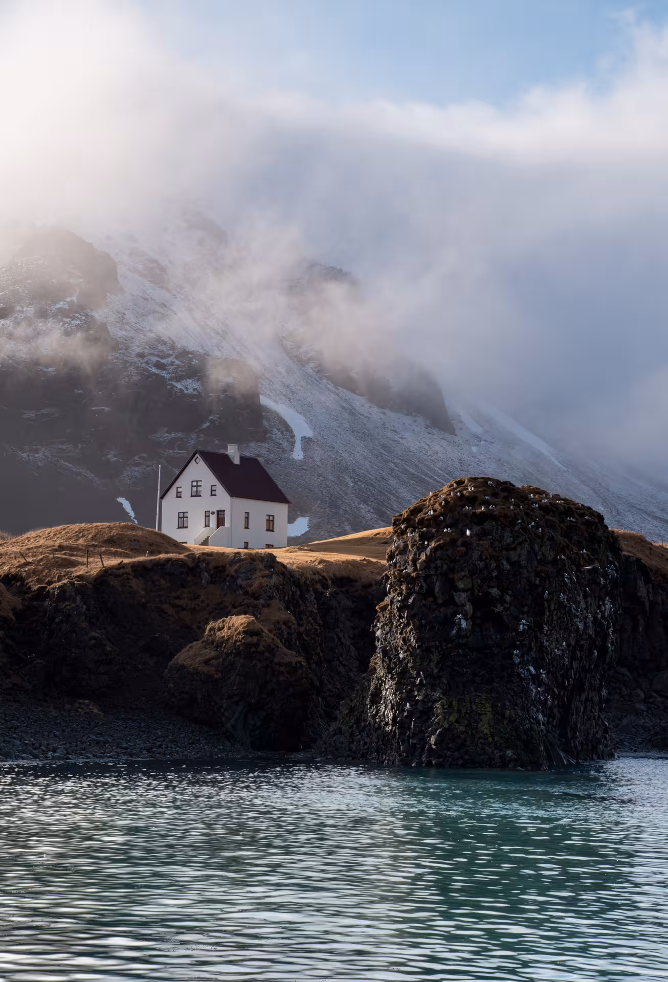 White house with red roof on rocky Snaefellsnes Peninsula coast with snow-capped mountain and mist behind